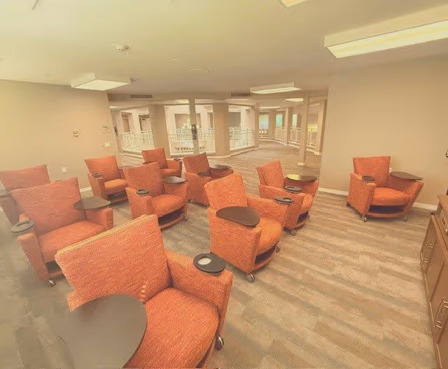 A spacious interior common area with multiple orange upholstered armchairs arranged in rows, each equipped with a small attached table. The room has beige walls, large windows, and a carpeted floor with a striped pattern. The area appears well-lit with ceiling lights and has an open layout with visible railings and doorways in the background.