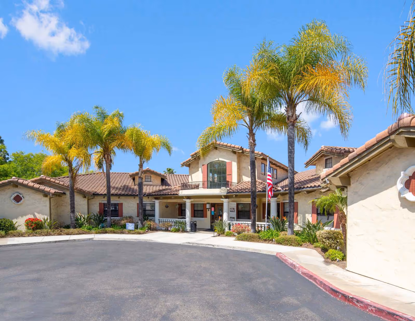 Exterior view of Grossmont Gardens Memory Care facility featuring a beige stucco building with red shutters and a tiled roof, surrounded by palm trees and landscaping under a clear blue sky.
