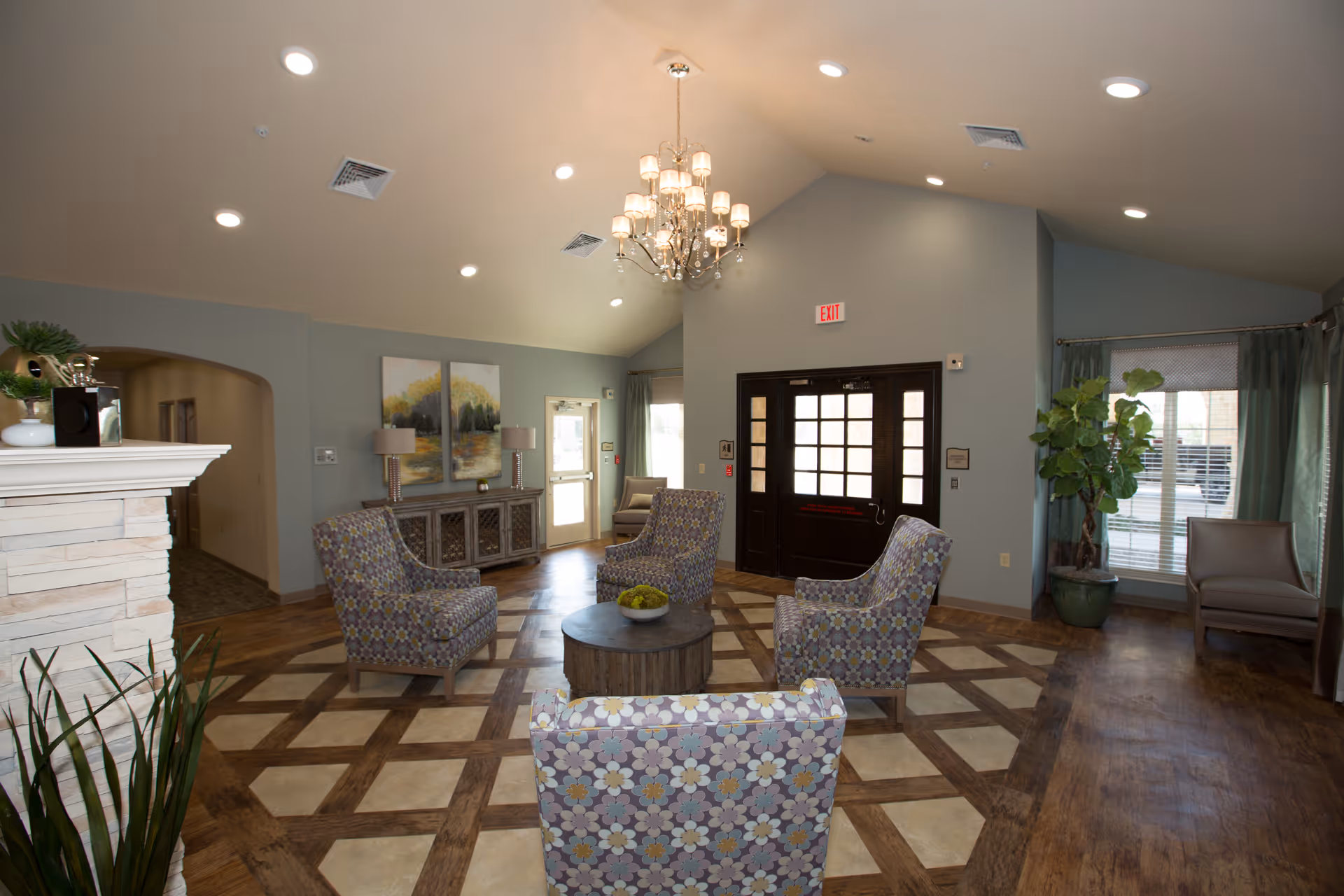 A spacious and well-lit common area with a high ceiling and a chandelier. The room features a patterned floor with a mix of wood and tile, four upholstered armchairs arranged around a round wooden coffee table with a bowl of green apples, a sideboard with two lamps and artwork above it, large windows with curtains, and a dark wooden door with glass panels. There is also a potted plant near the window and a fireplace partially visible on the left.