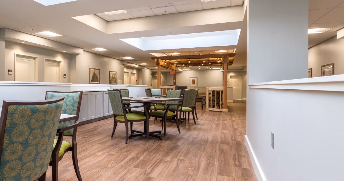 A spacious interior dining area with wooden flooring, several tables and chairs with green patterned upholstery, white walls, and ceiling skylights. The room is well-lit with recessed lighting and features a wooden pergola structure with string lights in the background.