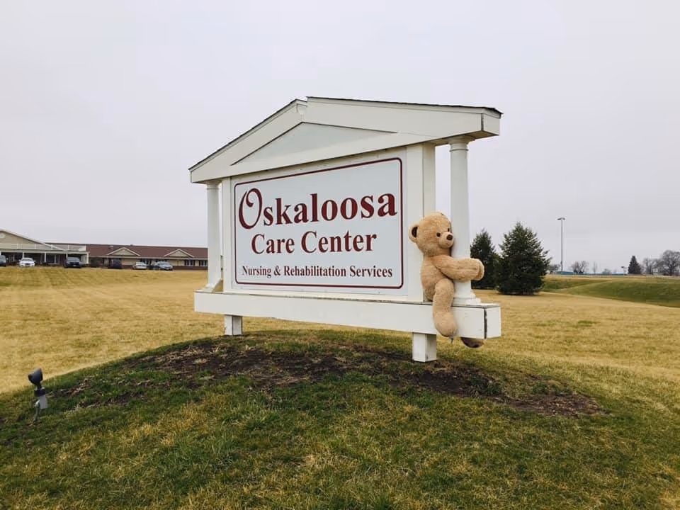 A large white sign reading "Oskaloosa Care Center Nursing & Rehabilitation Services" stands on a grassy lawn with a teddy bear attached and the facility building in the background.