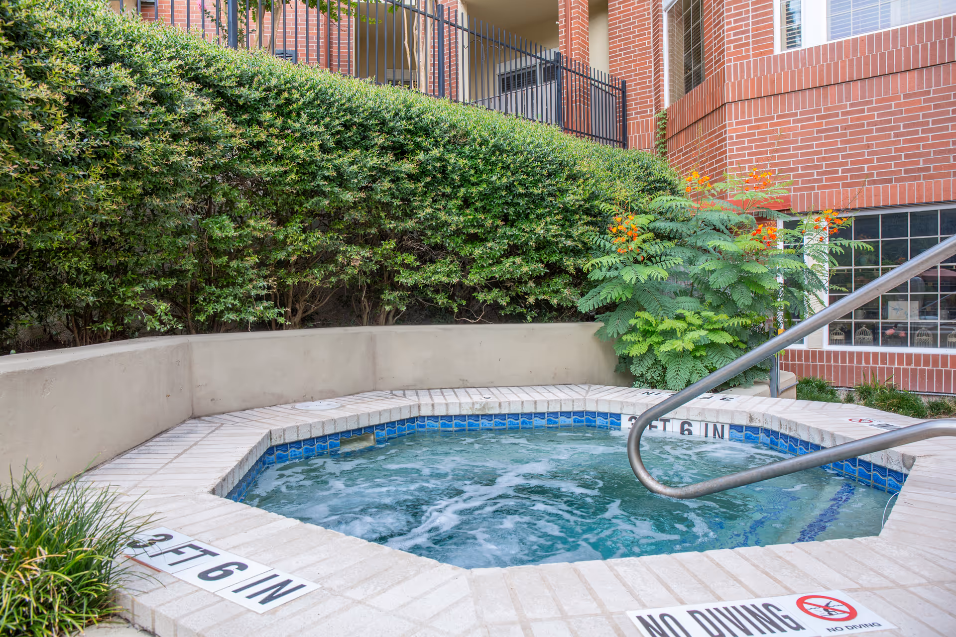 Outdoor hot tub or spa surrounded by a tiled edge with depth markings and a 'No Diving' sign, set against a backdrop of green bushes and a brick building with windows and a black metal fence.
