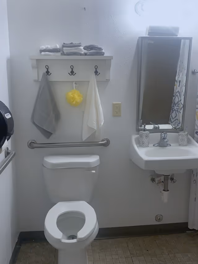 A small bathroom with a white toilet and a white wall-mounted sink. Above the toilet is a white shelf with three black hooks holding two towels and a yellow bath sponge. Several folded towels are stacked on the shelf. A mirror is mounted above the sink, and a soap dispenser and a small container are on the sink. The floor has small square tiles, and there is a metal grab bar on the wall behind the toilet.