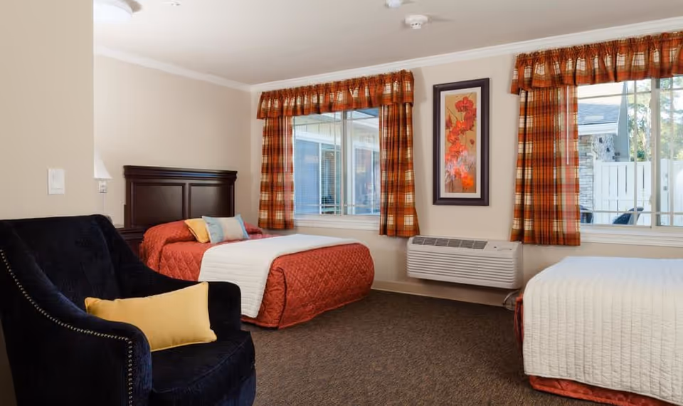 A cozy bedroom with two beds, one with a red quilt and the other with a white quilt. Both beds have pillows. There is a dark blue armchair with a yellow pillow in the foreground. The room has two windows with red and orange plaid curtains, a framed floral artwork on the wall, and a wall-mounted air conditioning unit below the artwork.