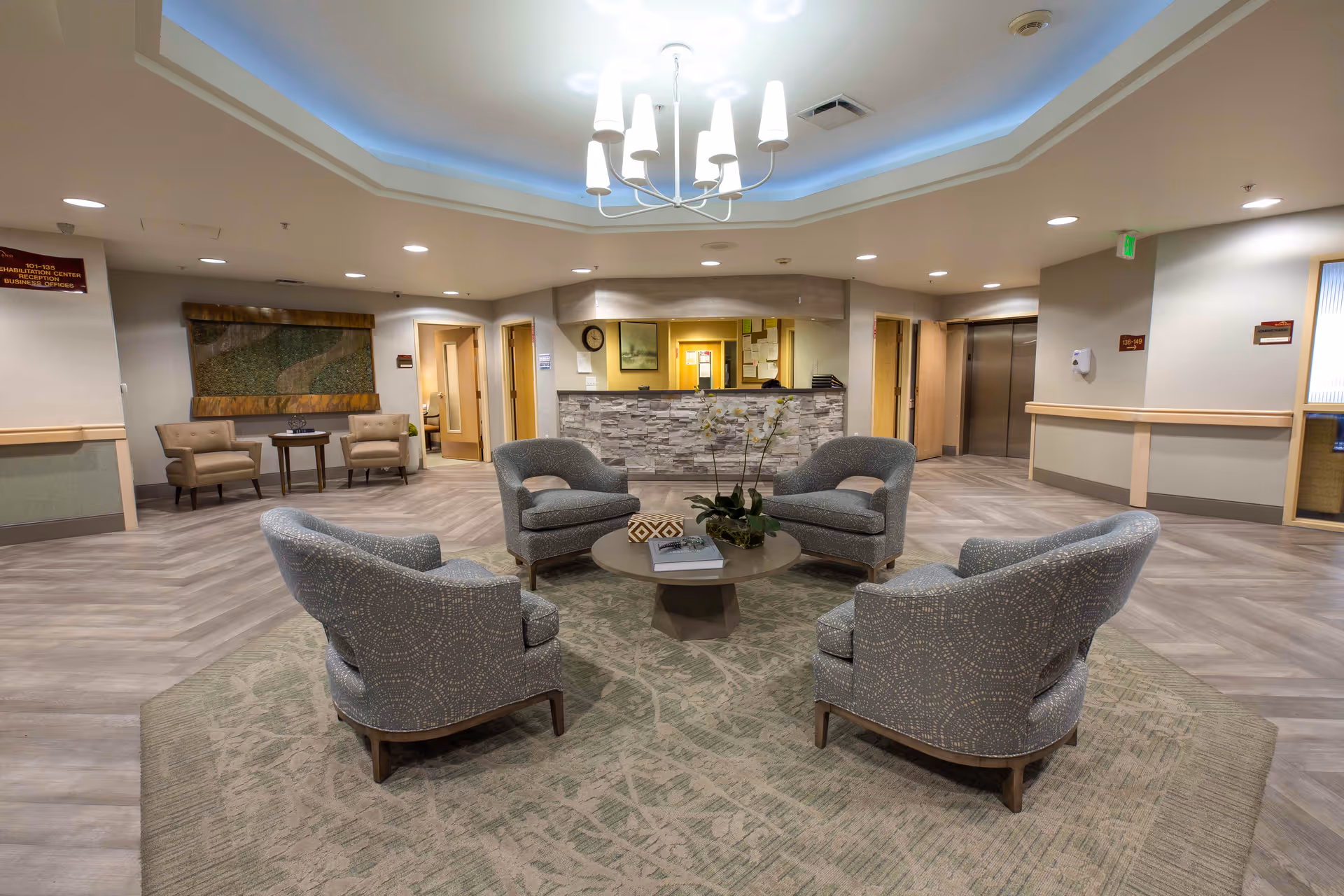 A spacious and well-lit reception area in Highland Care Center featuring a circular arrangement of five upholstered armchairs around a round coffee table with a plant and books. The floor has a patterned carpet under the seating area and wood-like flooring elsewhere. The reception desk with a stone facade is visible in the background, along with several doorways and an elevator. A modern chandelier hangs from the ceiling.