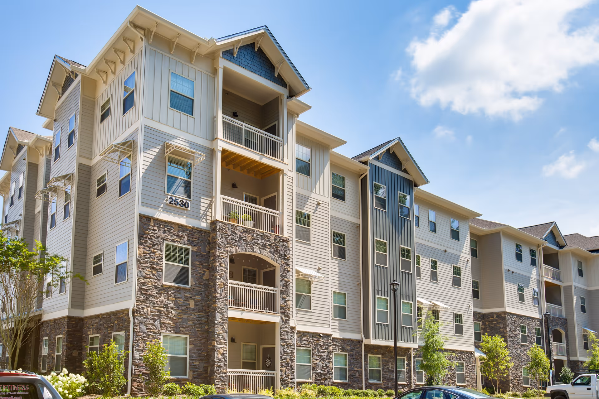 Exterior view of a modern multi-story residential building with stone and siding facade, balconies, and multiple windows under a partly cloudy blue sky.