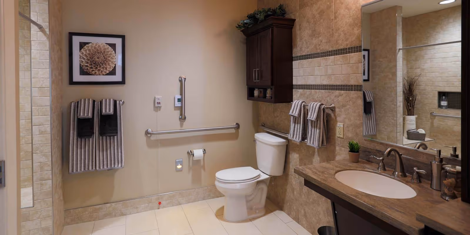 Accessible bathroom featuring a toilet with grab bars, a vanity sink with mirror, tiled walls and neatly hung towels.