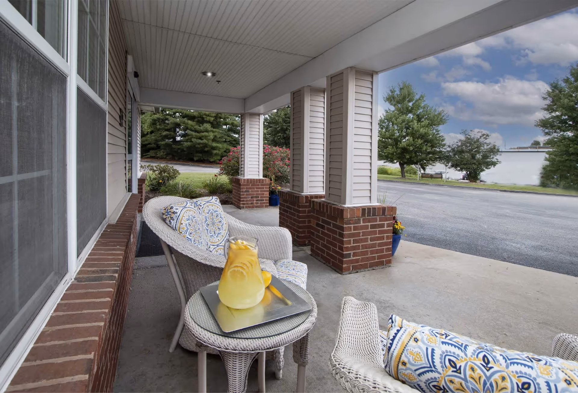 Covered outdoor patio area with white wicker chairs featuring patterned cushions and a small round table holding a pitcher of lemonade and a glass. The patio has brick and white siding columns, with a view of a parking area and trees in the background under a partly cloudy sky.