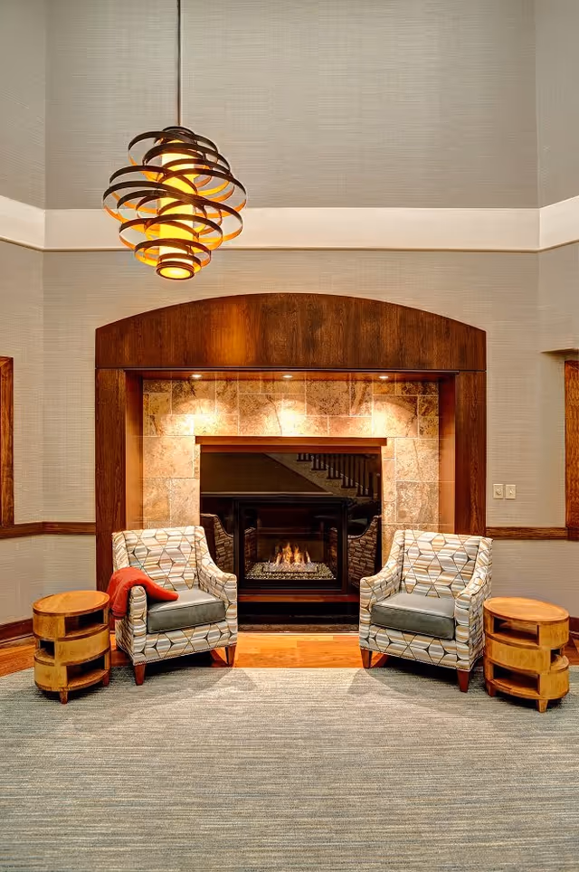 A cozy seating area with two patterned armchairs facing a lit fireplace framed by a wooden mantel and tiled surround. Each chair has a small wooden side table next to it, and one chair has a red throw draped over the arm. Above, a modern, circular pendant light hangs from the ceiling. The room features neutral-colored walls and a carpeted floor.