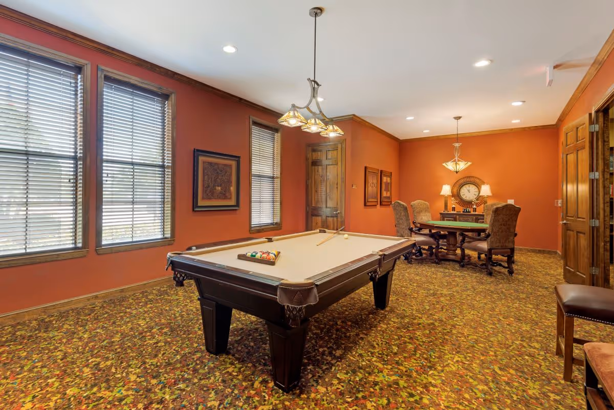 Game room with a pool table in the center, a card table and chairs in the background, warm orange walls and large windows.