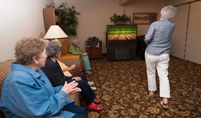 A group of elderly women in a senior living facility sitting on couches and chairs, watching another woman who is standing and playing a bowling video game on a large TV in a cozy room with patterned carpet and a lamp.