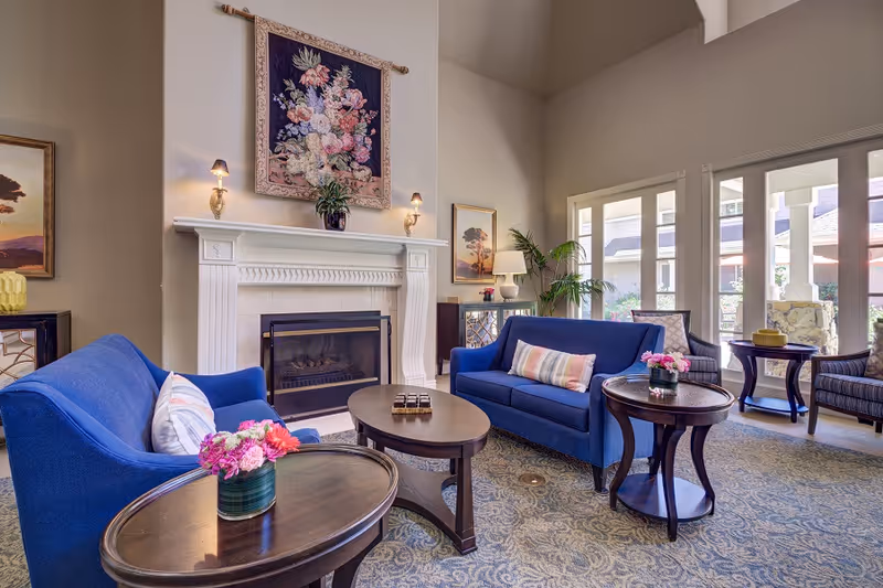 A cozy living room area with two blue sofas facing each other, a fireplace with a floral tapestry above it, and several wooden tables with flower arrangements. Large windows let in natural light and offer a view of an outdoor patio area.