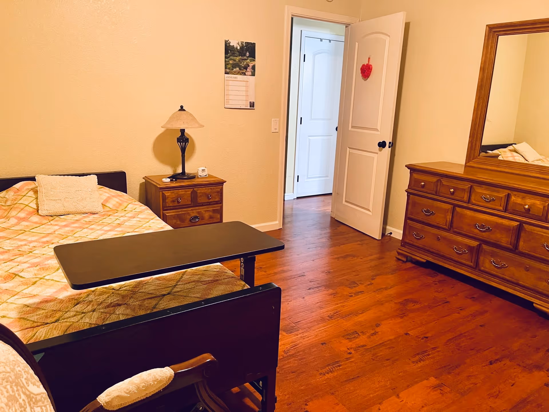 A senior living bedroom with a single bed covered in a patterned quilt, a wooden nightstand with a lamp and a small clock, a wooden dresser with a large mirror, and an open door leading to another room. The floor is wooden and the walls are painted beige.