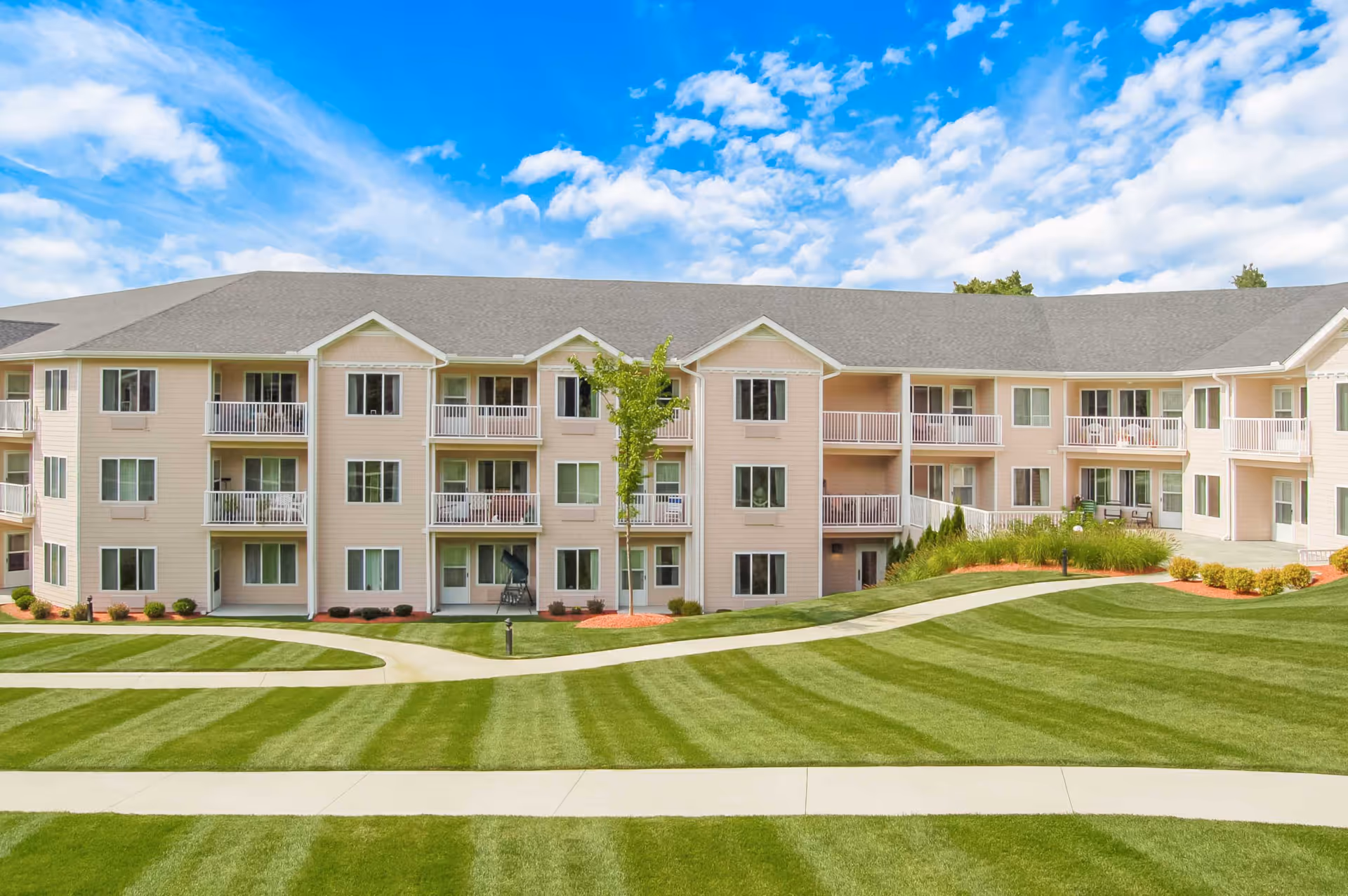 Exterior view of a three-story senior living facility building with beige siding and multiple balconies. The building is surrounded by well-maintained green lawns with striped mowing patterns and paved walkways. The sky is blue with scattered white clouds.
