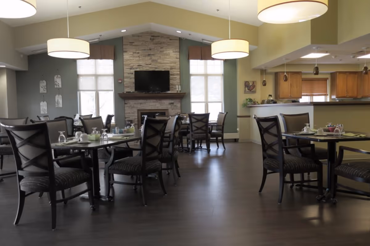 A dining area in a senior living facility with several tables and chairs arranged neatly. The room features a stone fireplace with a mounted flat-screen TV above it, large windows with blinds, and pendant lights hanging from the ceiling. The space has a warm and inviting atmosphere with a view into a kitchen area in the background.