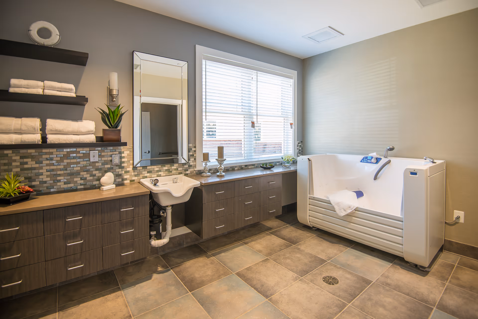 A modern bathroom with a walk-in bathtub on the right side, a small white sink with a mirror above it in the center, and wooden cabinets with drawers and shelves holding neatly folded white towels and decorative plants. The floor is tiled, and there is a large window with blinds letting in natural light.