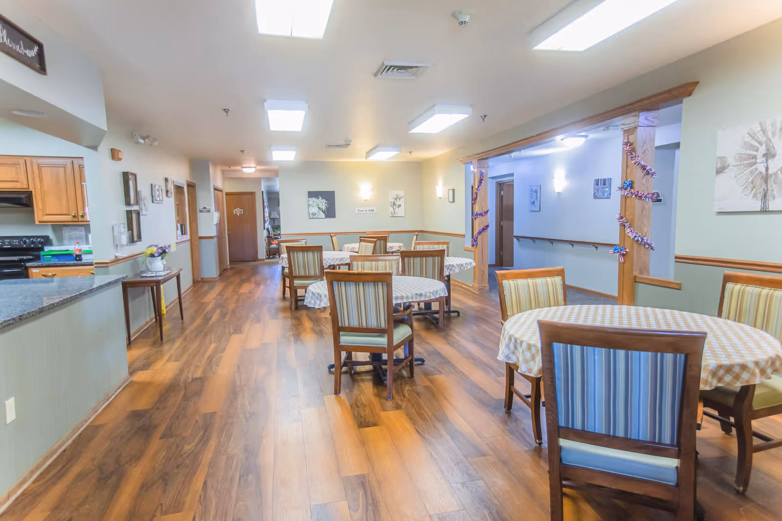 A bright dining area in a senior living facility with several round tables covered with checkered tablecloths and wooden chairs with striped cushions. The room has wooden flooring, light-colored walls, and ceiling lights. There is a kitchen area visible on the left side with wooden cabinets and a black stove. The right side has a hallway decorated with garlands.