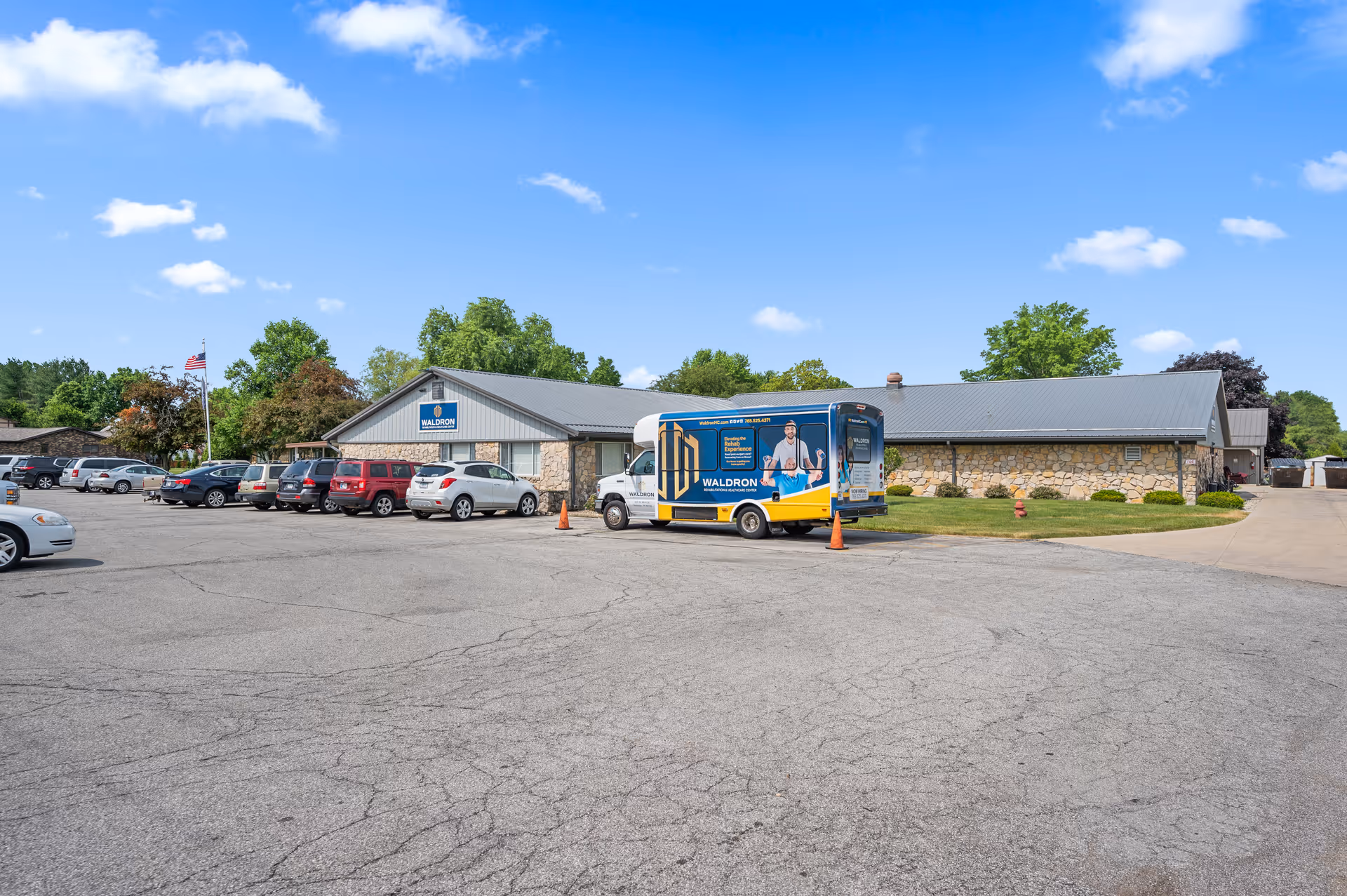 Exterior view of Waldron Rehabilitation and Healthcare Center building with a parking lot in front. Several cars are parked, and a branded Waldron vehicle is visible. The building has a stone facade and a metal roof, with trees and a clear blue sky in the background.