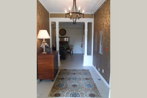 Narrow furnished interior hallway with a patterned rug, wooden side table and lamp, wallpapered walls, and a chandelier leading into a living area.