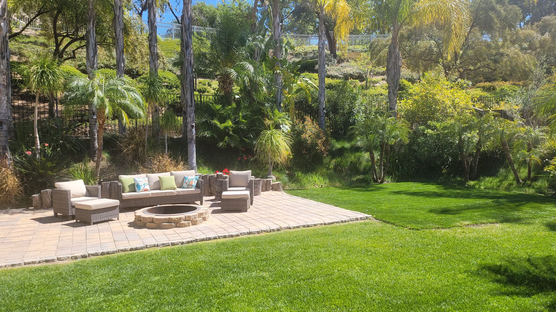 Outdoor patio area with cushioned wicker seating arranged around a circular stone fire pit, surrounded by lush green grass, palm trees, and various other plants and shrubs under a sunny sky.