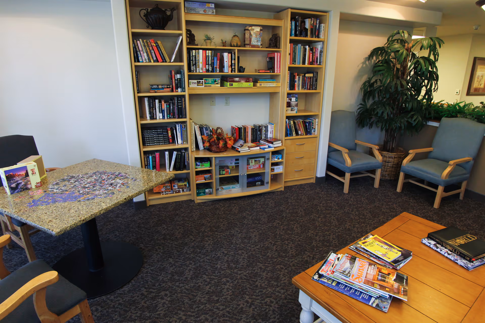 A cozy common area with a bookshelf filled with books and board games, two blue cushioned chairs, a table with magazines and books, and another table with a partially completed jigsaw puzzle. There is a large potted plant in the corner and neutral-colored walls and carpet.