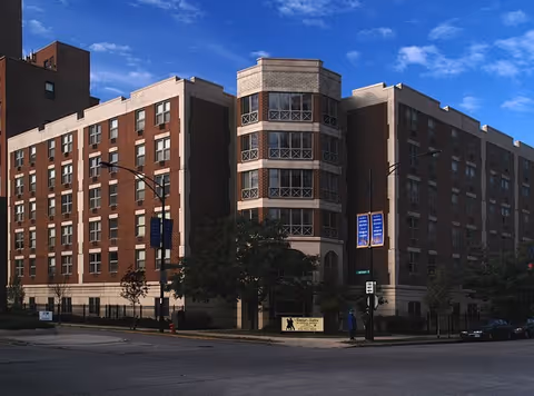 Brick multi-story senior living building on a street corner under a blue sky.