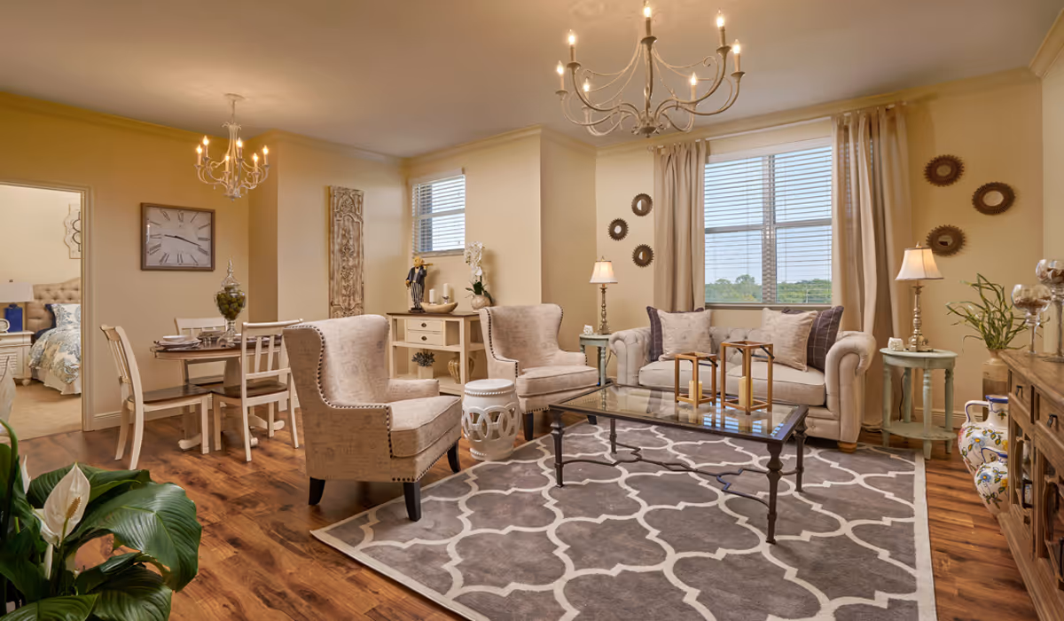 Sunlit living room with beige sofas and armchairs around a glass coffee table, a patterned rug, adjacent dining area and chandeliers.