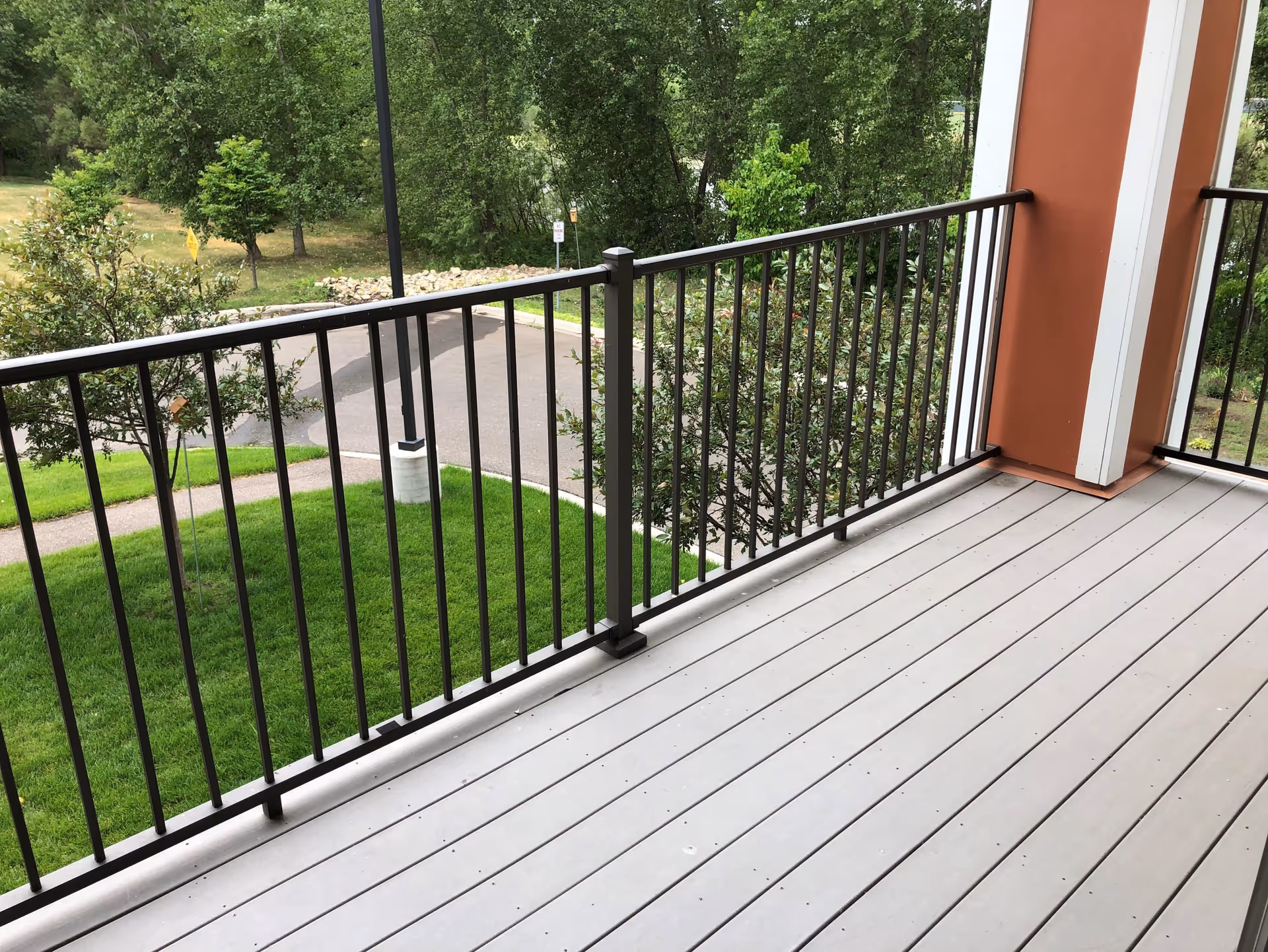 View from a balcony with gray wooden flooring and black metal railing overlooking a green lawn, trees, and a paved road with a lamppost.