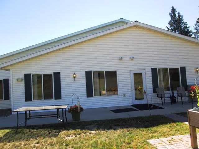 Exterior view of a single-story building with white siding and green shutters. There are three windows and a door with glass panels. Outside, there is a table, some chairs, potted plants, and a grassy area in front of the building under a clear sky.