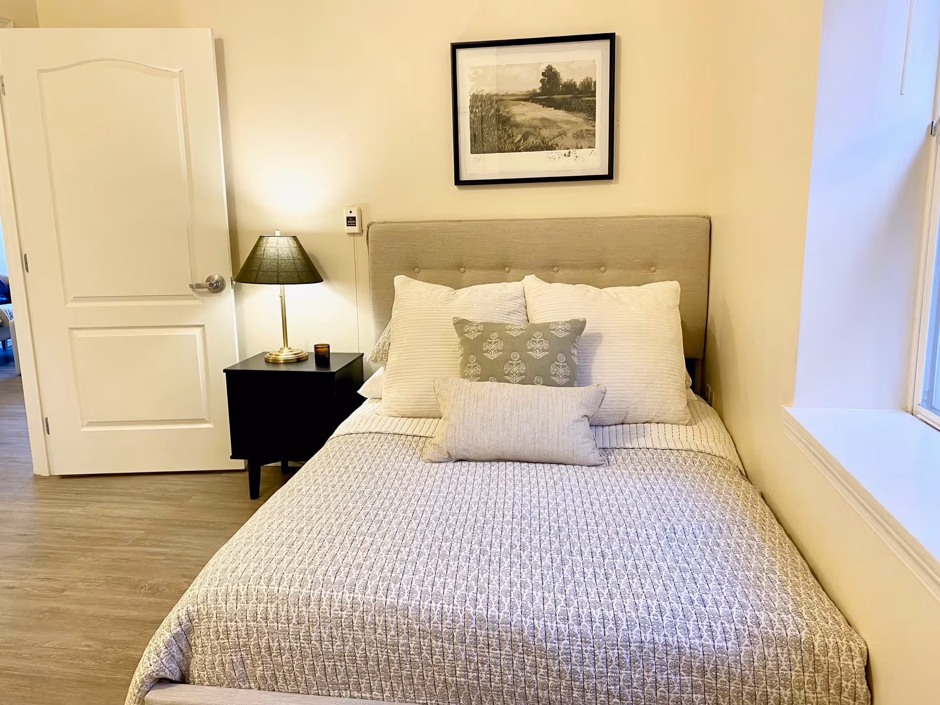 A neatly made bed with multiple pillows in a bedroom. There is a black nightstand with a lamp and a small candle on it next to the bed. A framed landscape artwork hangs on the wall above the headboard. The room has light-colored walls and wood flooring, with a partially open white door and a window with a wide sill.