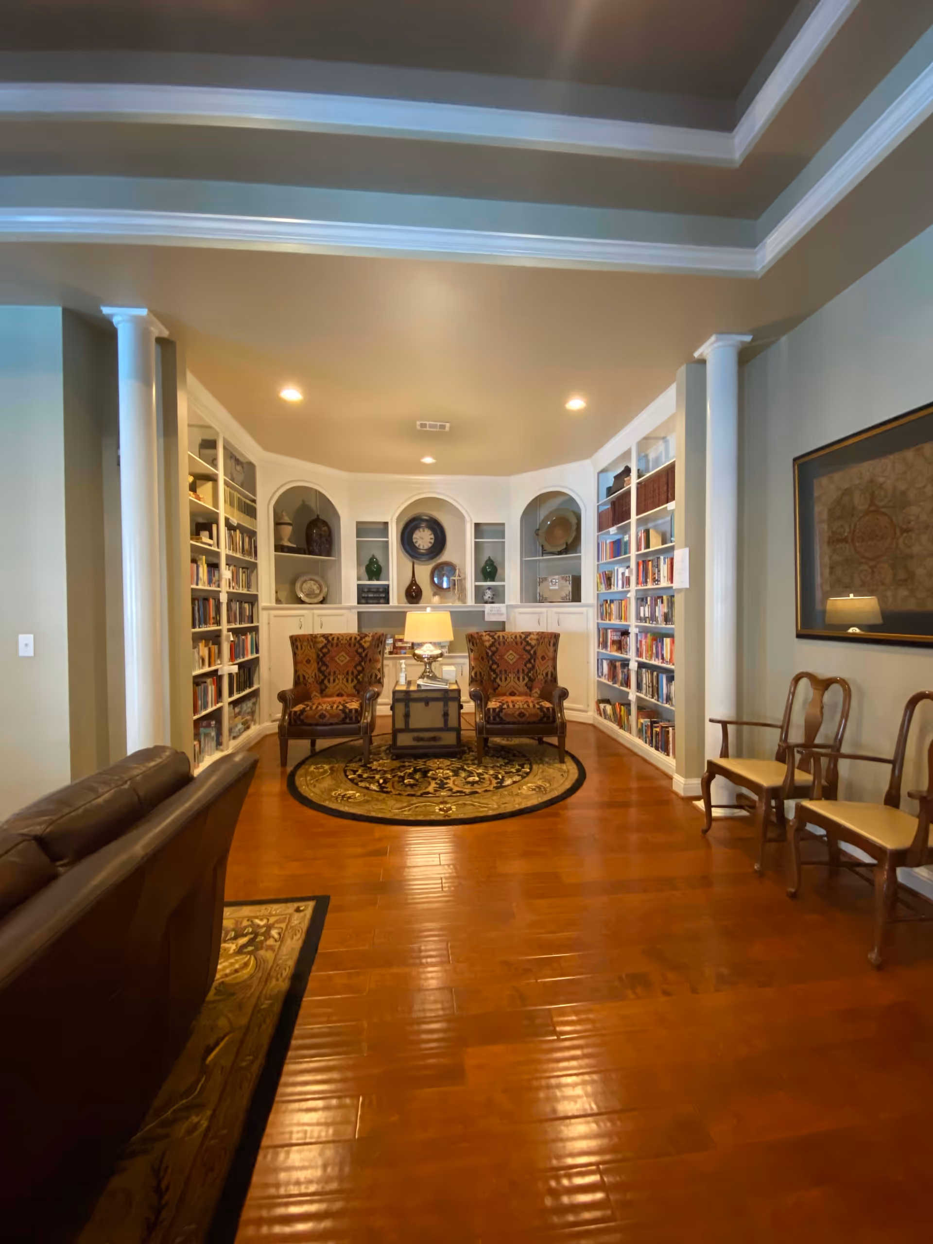A cozy library or reading room with wooden floors, two patterned armchairs placed on a round decorative rug, a small table with a lamp between them, built-in white bookshelves filled with books and decorative items, and additional wooden chairs along the right wall. The ceiling has recessed lighting and crown molding.