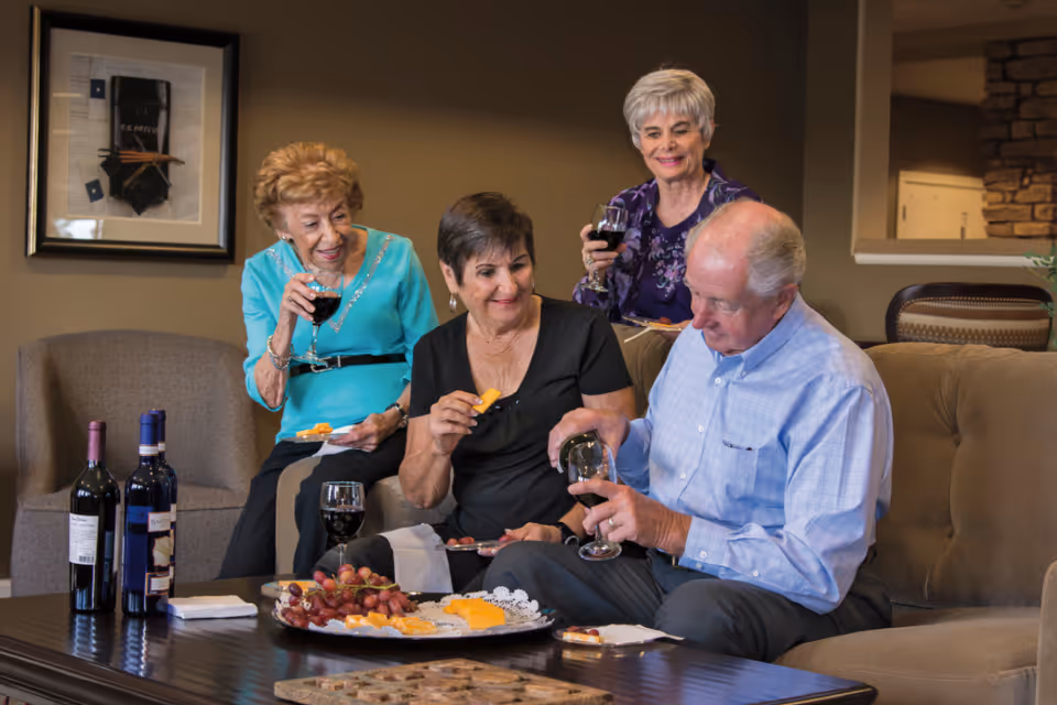Four older adults sitting in a living room lounge sharing wine and cheese from a platter.