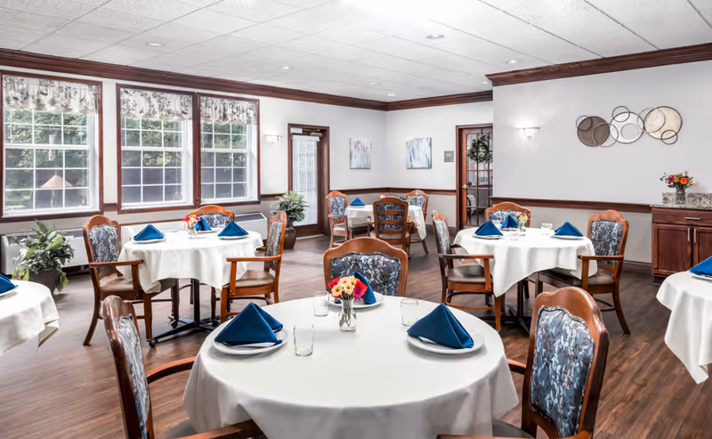 A bright dining room with round tables covered in white tablecloths, each set with blue folded napkins, plates, glasses, and small flower arrangements. The room has large windows with floral valances, wooden chairs with patterned upholstery, wood flooring, and light-colored walls with decorative artwork and plants.