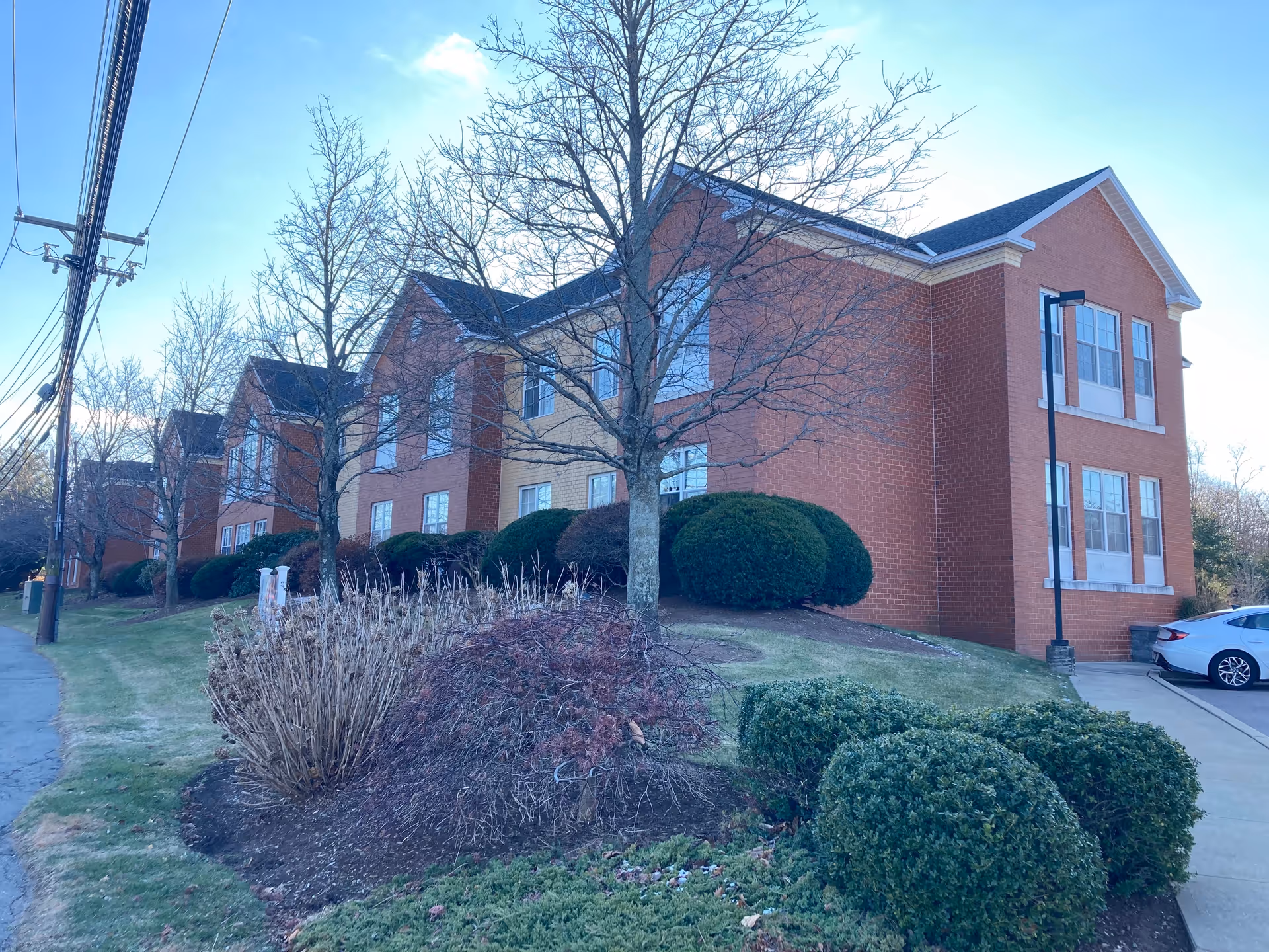 Exterior view of a multi-story brick building with large windows, surrounded by trimmed bushes, leafless trees, and a sidewalk. A white car is parked on the right side near the building.