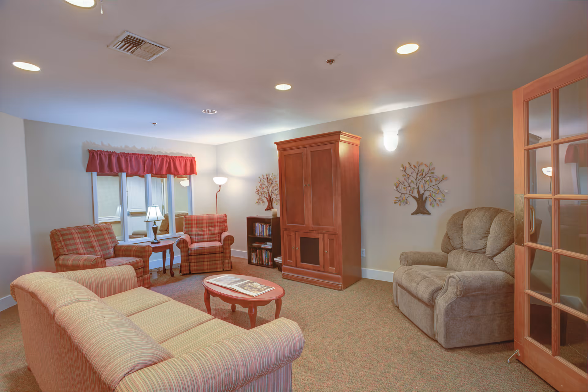 A cozy living room with a striped sofa, two plaid armchairs, a cushioned recliner, a wooden coffee table with magazines, a wooden cabinet, a small bookshelf, and a floor lamp. The room has beige walls, carpeted floor, and a window with a red valance. There is a decorative metal tree wall art and a wooden door with glass panels.