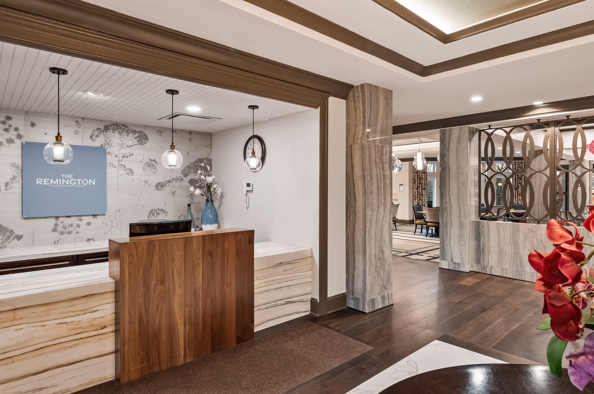 Reception area of The Remington of McCandless featuring a wooden front desk with marble accents, two hanging pendant lights, a wall clock, and a decorative vase with flowers. The background includes a wall with floral patterns and a sign that reads 'The Remington'. To the right, there is a glimpse of a seating area with chairs and a decorative partition.