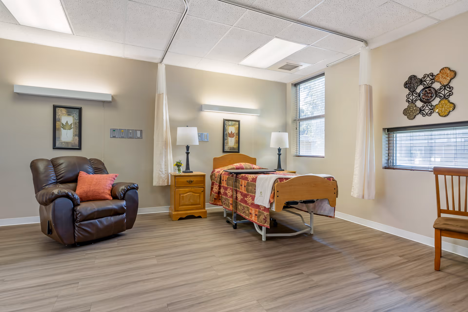A well-lit room in a senior living facility featuring a single hospital-style bed with a patterned bedspread, a wooden nightstand with a lamp and small plant, a comfortable brown recliner chair with a red cushion, a wooden chair, and decorative wall art. The room has large windows with blinds and curtains, and wood-look flooring.