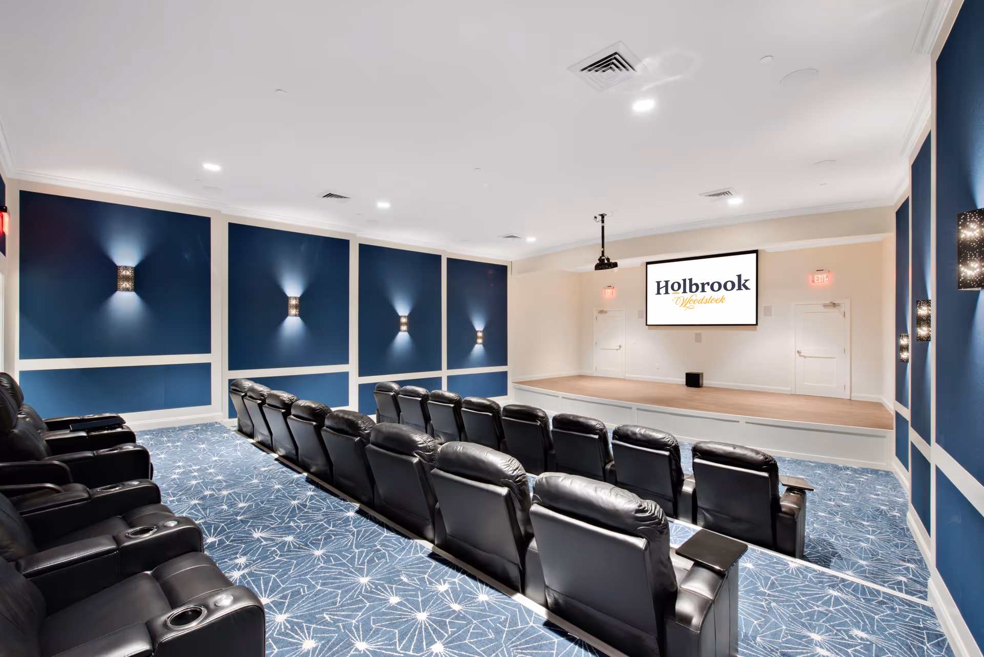 Small screening room with rows of black leather recliners facing a stage and a screen displaying 'Holbrook Woodstock'.