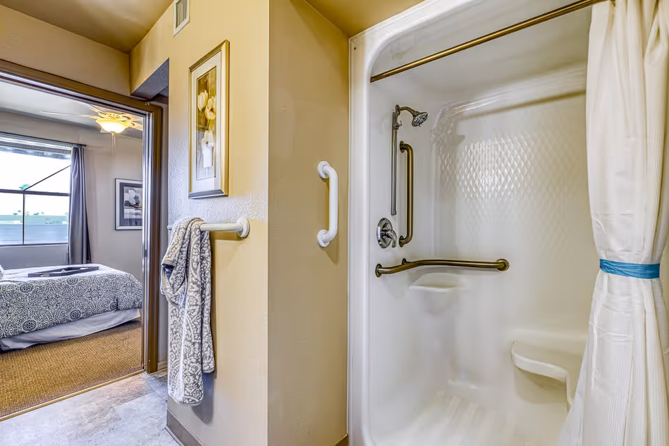 View of a bathroom shower with grab bars and a white shower curtain tied back, adjacent to a bedroom with a bed covered in a patterned bedspread and a ceiling fan above.