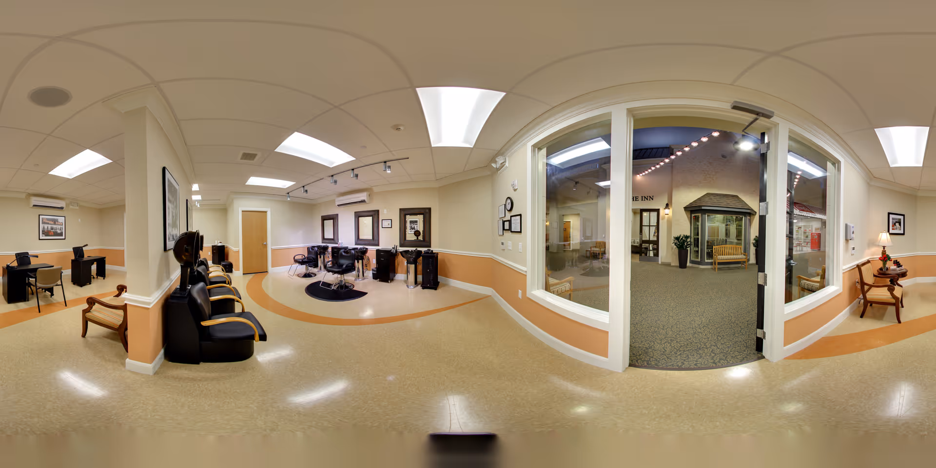 Interior view of a senior living facility's common area with a hair salon section featuring salon chairs and mirrors, a seating area with chairs and a small table with a lamp, and a large windowed door leading to an indoor courtyard or lobby area with benches and decorative lighting.