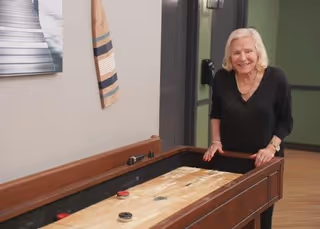 An elderly woman with white hair, wearing a black top, stands smiling next to a wooden shuffleboard table in an indoor recreational area with light-colored walls and a piece of wall art.
