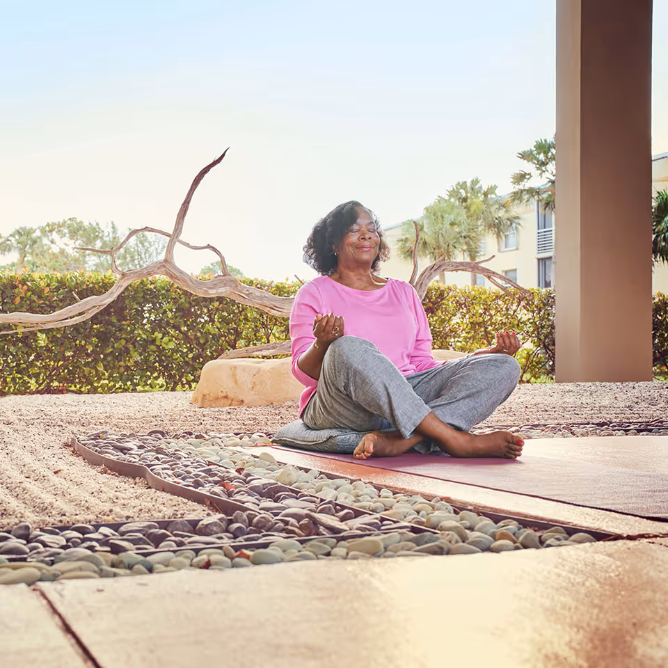 An elderly woman wearing a pink top and gray pants is sitting cross-legged on a yoga mat outdoors, meditating with her eyes closed and hands resting on her knees. Behind her is a hedge, some palm trees, and a building structure.