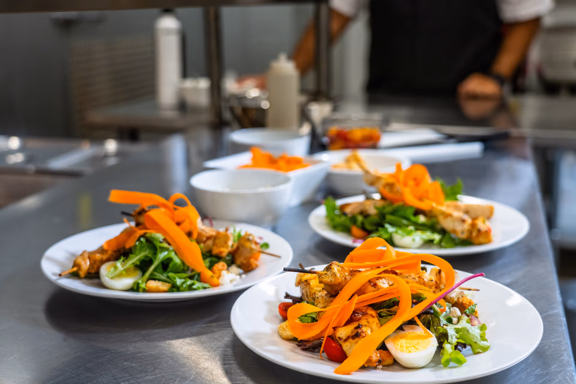 Three plates of fresh salad with grilled chicken skewers, leafy greens, halved boiled eggs, cherry tomatoes, and carrot ribbons arranged on a stainless steel kitchen counter with a blurred background of kitchen equipment and a person in an apron.