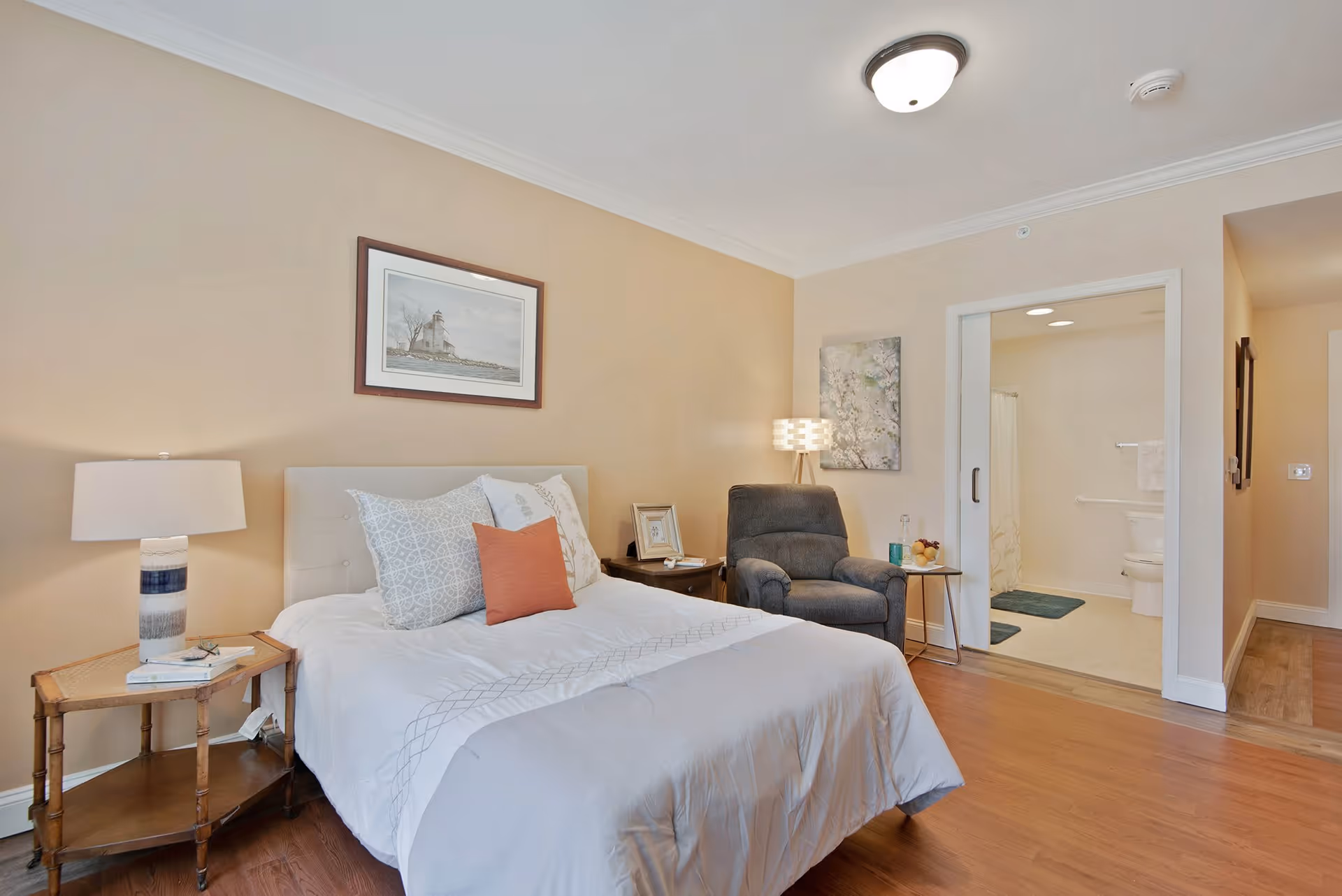 A cozy bedroom with a neatly made bed featuring white and patterned pillows and a coral accent pillow. There is a wooden side table with a lamp and books on the left side of the bed. A comfortable gray armchair with a small round side table holding a glass and a bowl of fruit is positioned near the entrance to a bathroom. The bathroom has a shower curtain, toilet, and blue bath mats. The walls are painted beige, and there is a framed picture of a lighthouse above the bed and a floral painting near the armchair. The floor is wooden, and the room is well-lit with ceiling lights.