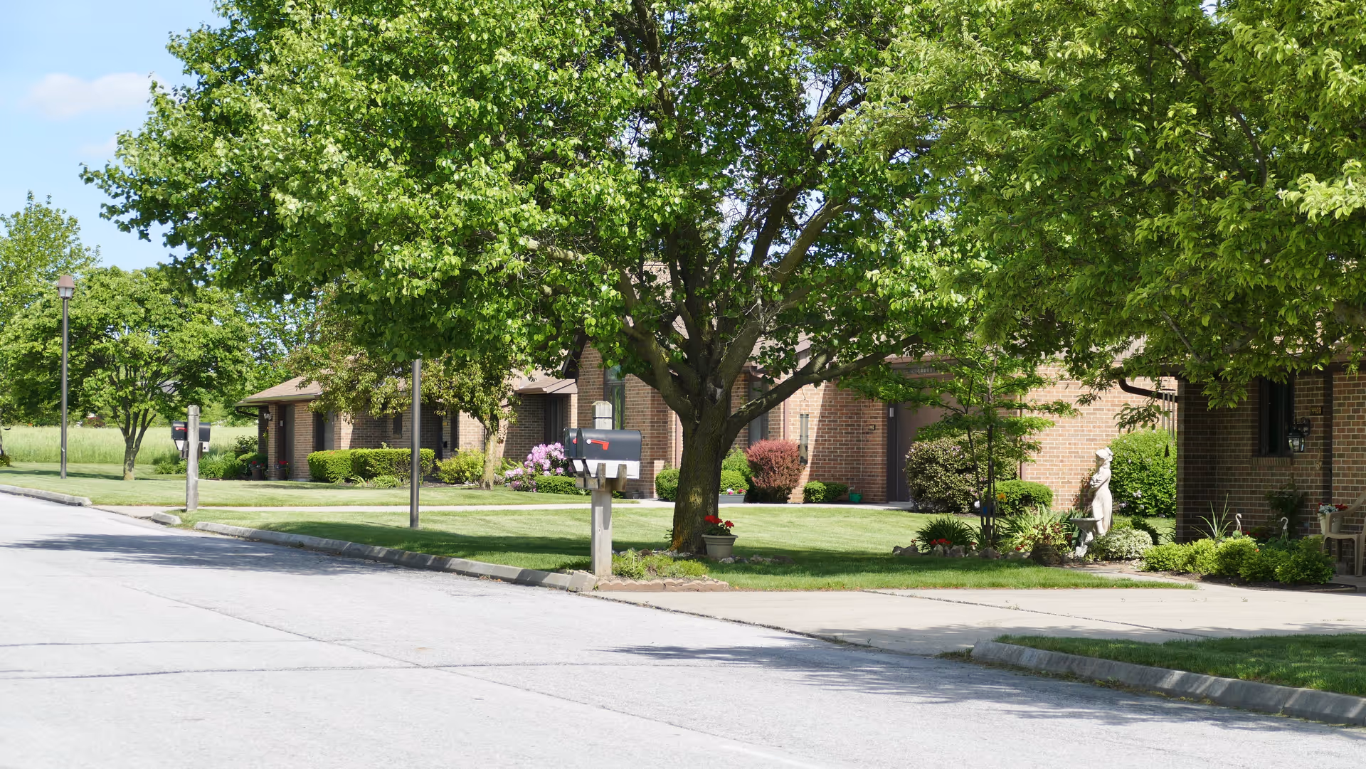 A quiet residential street in a senior living community with brick single-story buildings, green lawns, trees, and mailboxes along the sidewalk on a sunny day.