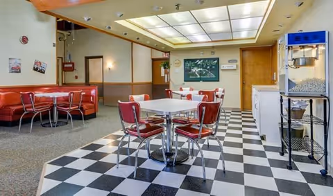 Dining area with black-and-white checkered floor, chrome tables with red chairs and a red booth, a popcorn machine, and a large lighted ceiling panel.