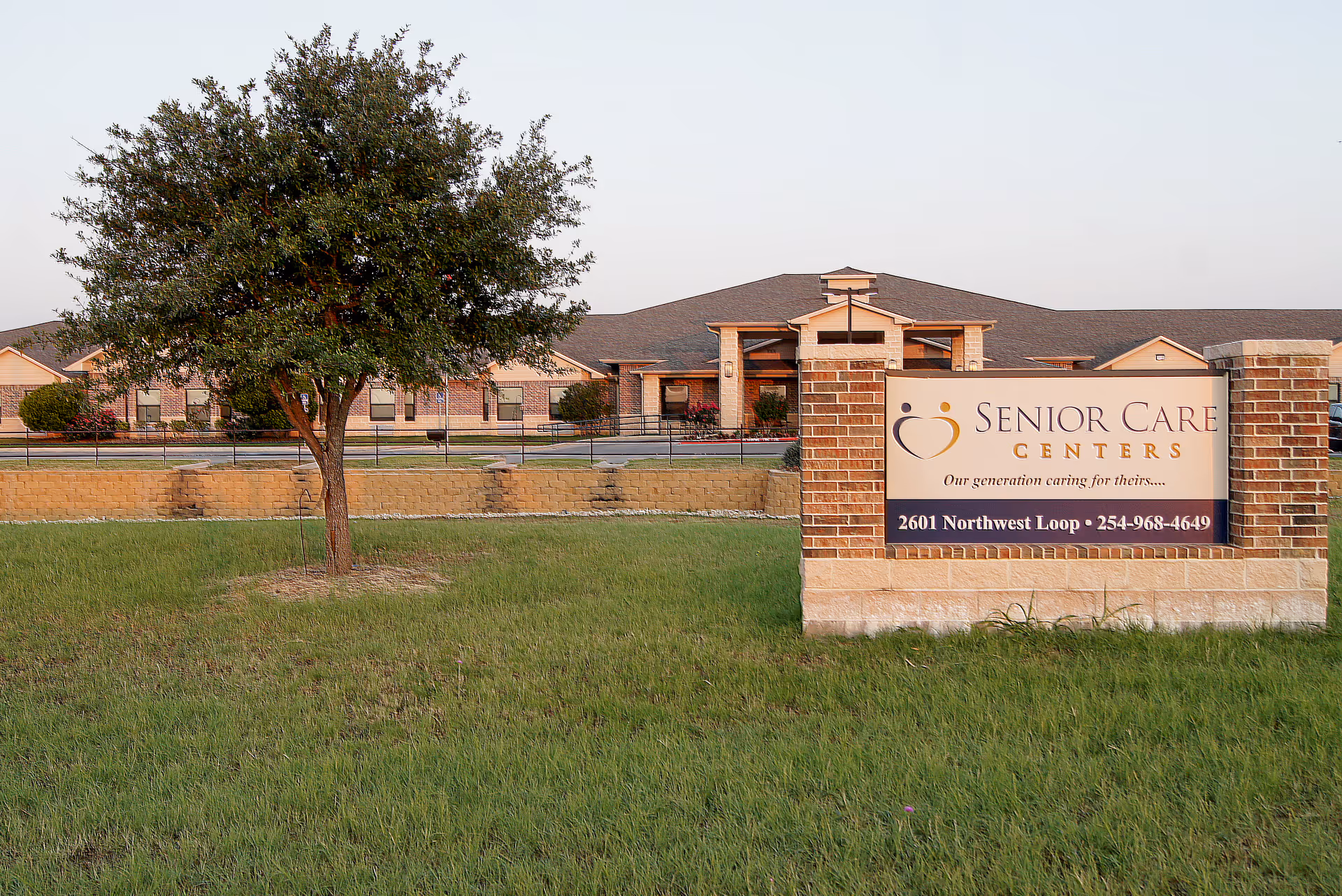 Exterior front view of a senior care center with a grassy lawn, a tree, and a brick sign reading 'Senior Care Centers'.