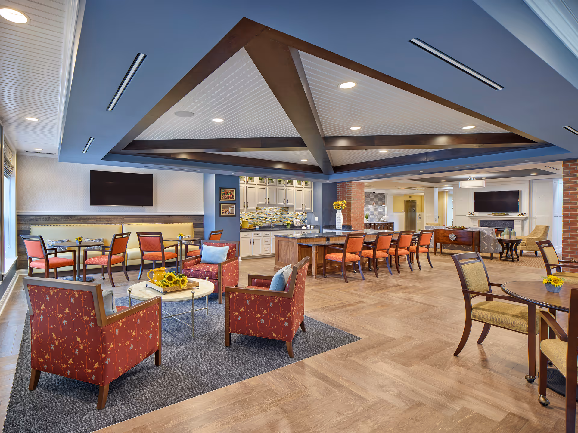 A spacious and well-lit common area in a senior living facility featuring multiple seating arrangements including red patterned armchairs around a small round table with sunflowers, a long counter with orange chairs, and several dining tables with chairs. The room has a decorative ceiling with wooden beams, a large flat-screen TV mounted on the wall, and a kitchen area with white cabinets and a tiled backsplash in the background.