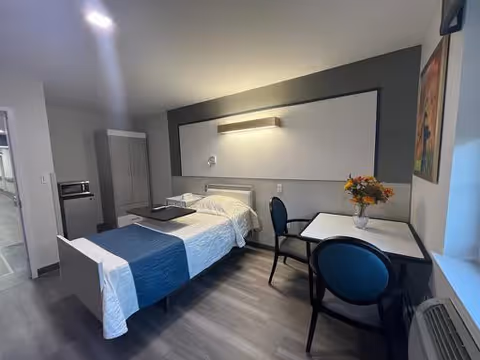 A clean and modern bedroom in a senior living facility featuring a single bed with white and blue bedding, a small table with two chairs, a vase with flowers on the table, a wardrobe, and a window with natural light coming in.