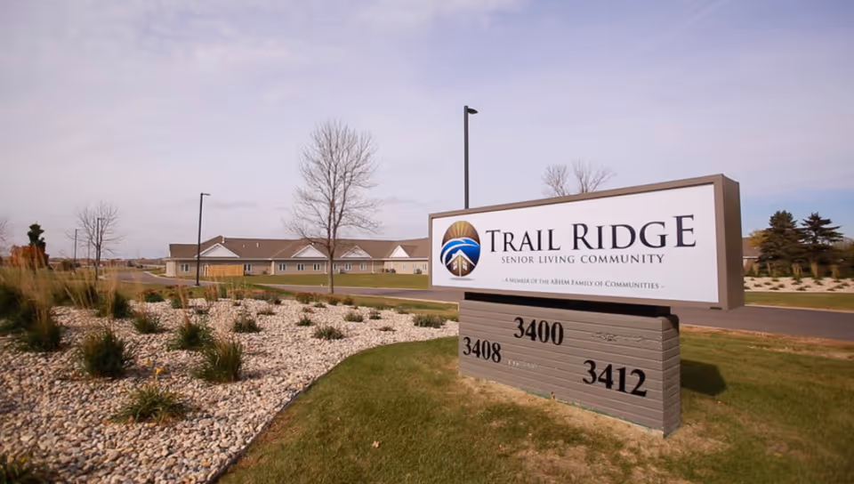 Outdoor view of the Trail Ridge Senior Living Community sign with a landscaped area of rocks and plants in the foreground and a single-story building in the background under a partly cloudy sky.